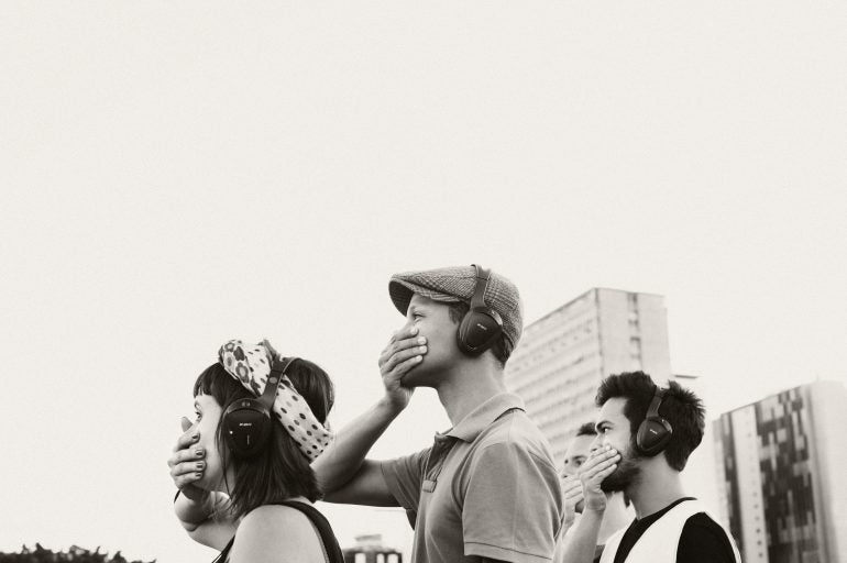 Image description: a woman and three men standing in profile, wearing headphones, all with their hands over their mouths.