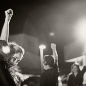 image description: in foreground: a woman, wearing headphones, with her fist in the air. In background: more people—all with headphones on—with their fists raised.