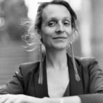 Professional black and white headshot of Emma Jaster, a woman with hair pulled back wearing dangling earrings and a dark blazer, smiling with arms crossed