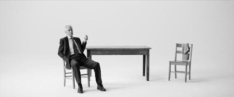 David Strathairn on stage white background with a chair and table