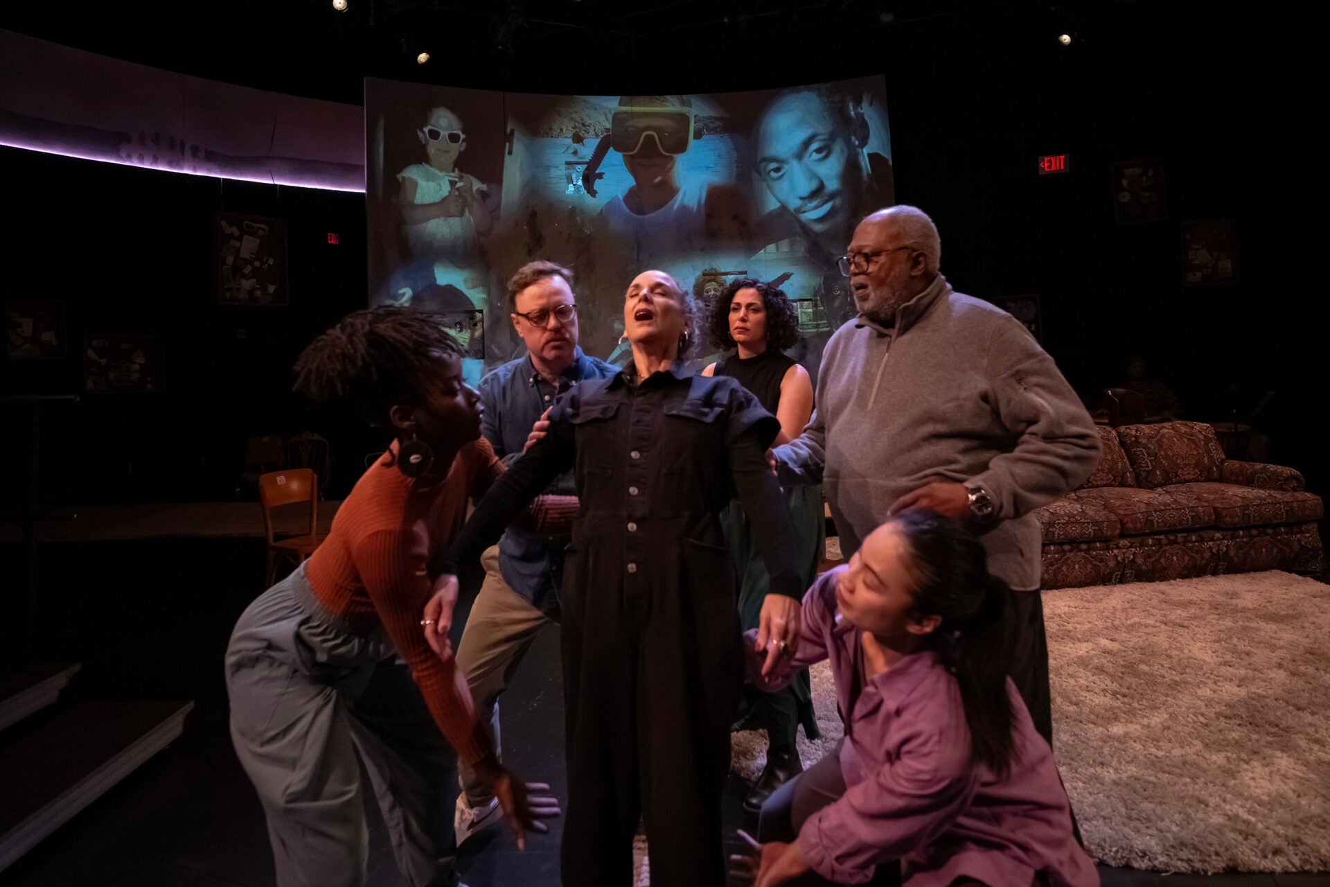 Woman in black jumpsuit surrounded by other men and women on a stage with a couch, rug, and screen in the background with memoriam images