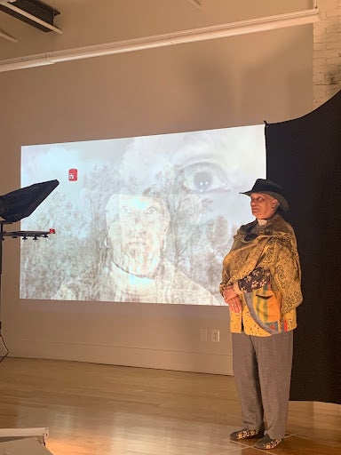 Man in a cowboy hat stands in a small rehearsal space in front of a screen showing self portraits