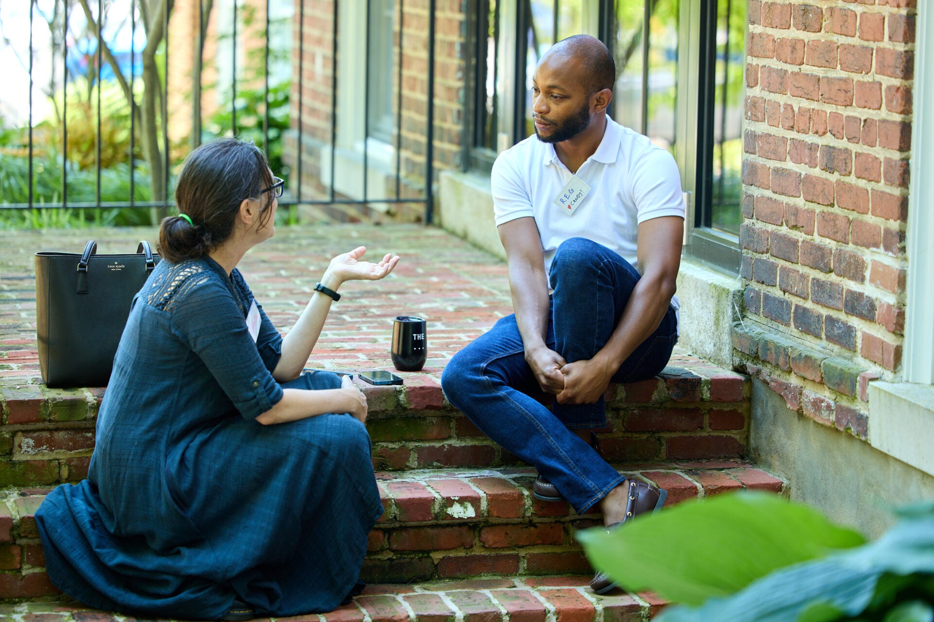 Man with shaved head, white polo shirt, and jeans sits on brick steps outdoors and listens to woman with dark bun and blue plaid dress who is speaking