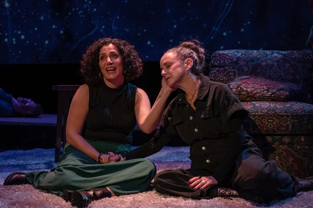 Woman with short curly dark hair sits on the rug beside a woman in a black jumpsuit, cups her face, and holds her hand