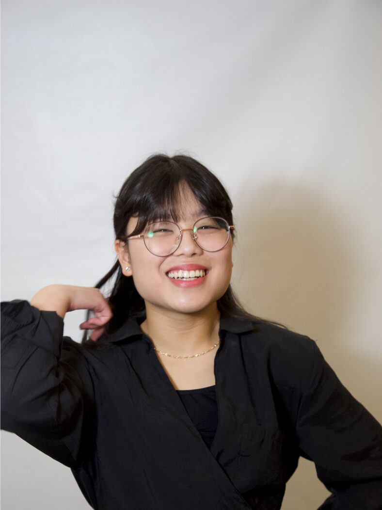 A young Asian woman with dark hair and glasses wearing a black outfit, smiling while posing with her hand near her face against a light background.
