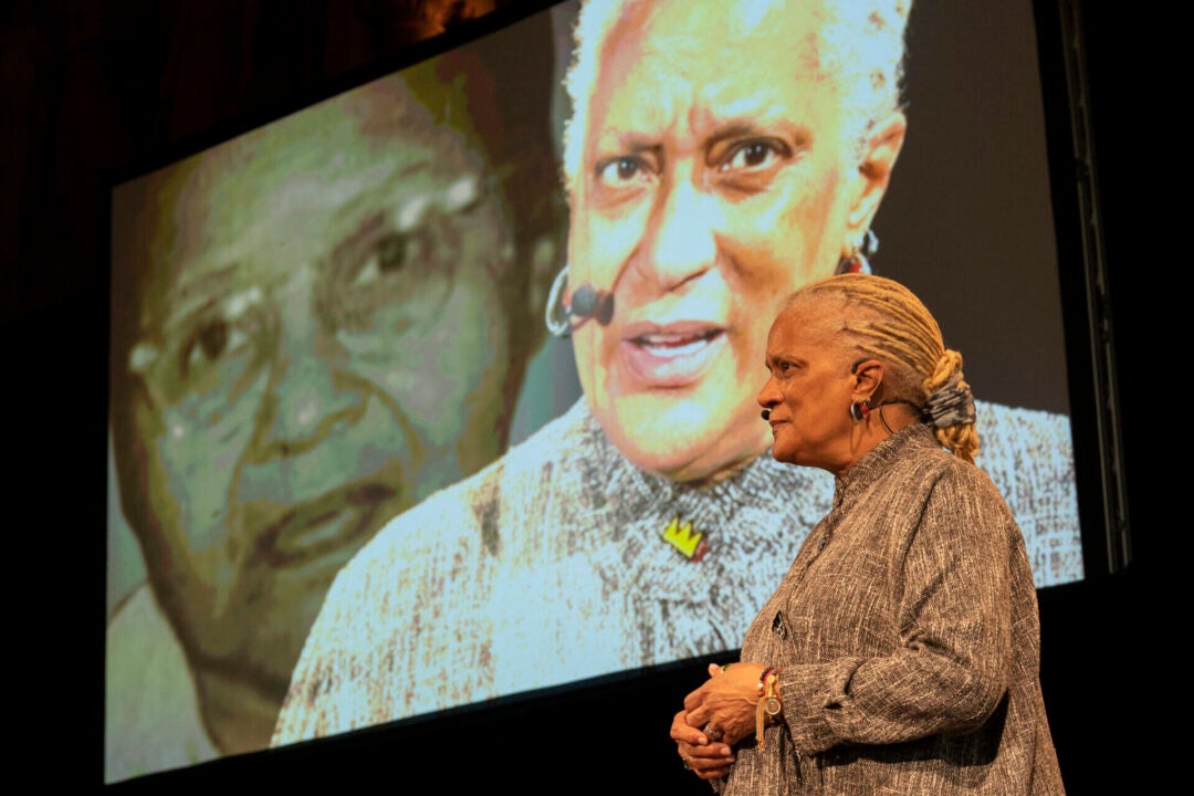 Older woman with blonde braided ponytail and gray dress stands in front of a screen that is capturing her speaking in real time and also showing an image of an older woman in glasses