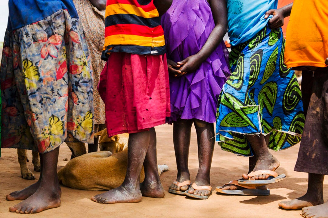 From the chest down, a line of children in colorful skirts standing on a dirt floor