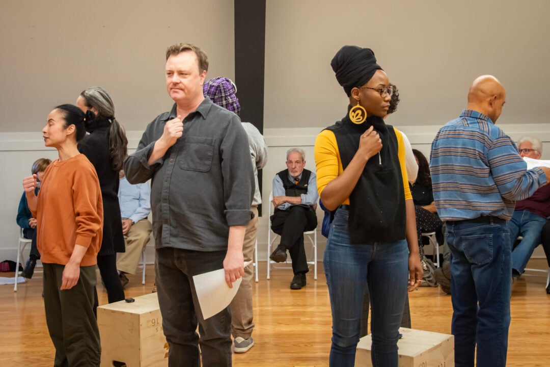 Group of men and women stand in a circle surrounded by watching audience members and face outwards with their closed fists against their chests