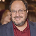 Professional headshot of Derek Goldman, a man with glasses, beard, and receding hairline wearing a dark blazer over a burgundy shirt, smiling at the camera
