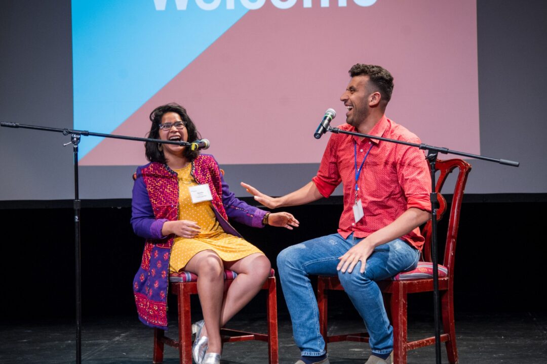 Dark haired man and dark haired woman sit in chairs on a stage behind microphones and laugh