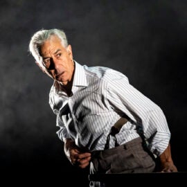 Older man with silver hair in a striped white button down poses dramatically in a dark space and looks off camera